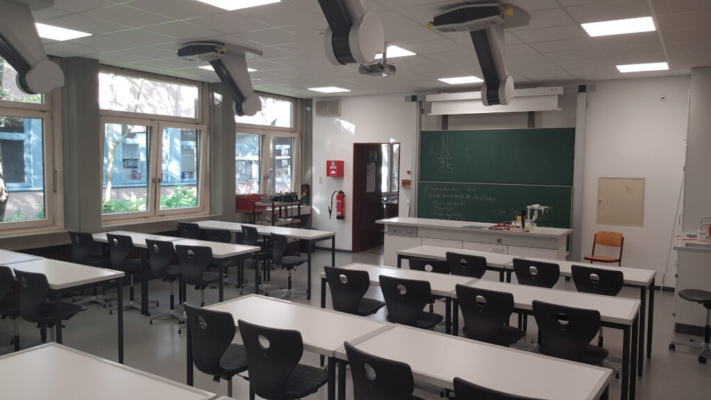 Raum mit Tischreihen und Stühlen dran. An der Decke sind Stationen mit Ansschlüssen für Gas und Strom, die für Experimente herunter gelassen werden können.